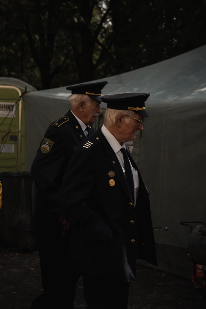 Dva starší hasiči ve slavnostních uniformách stojí u stanů během venkovní akce.