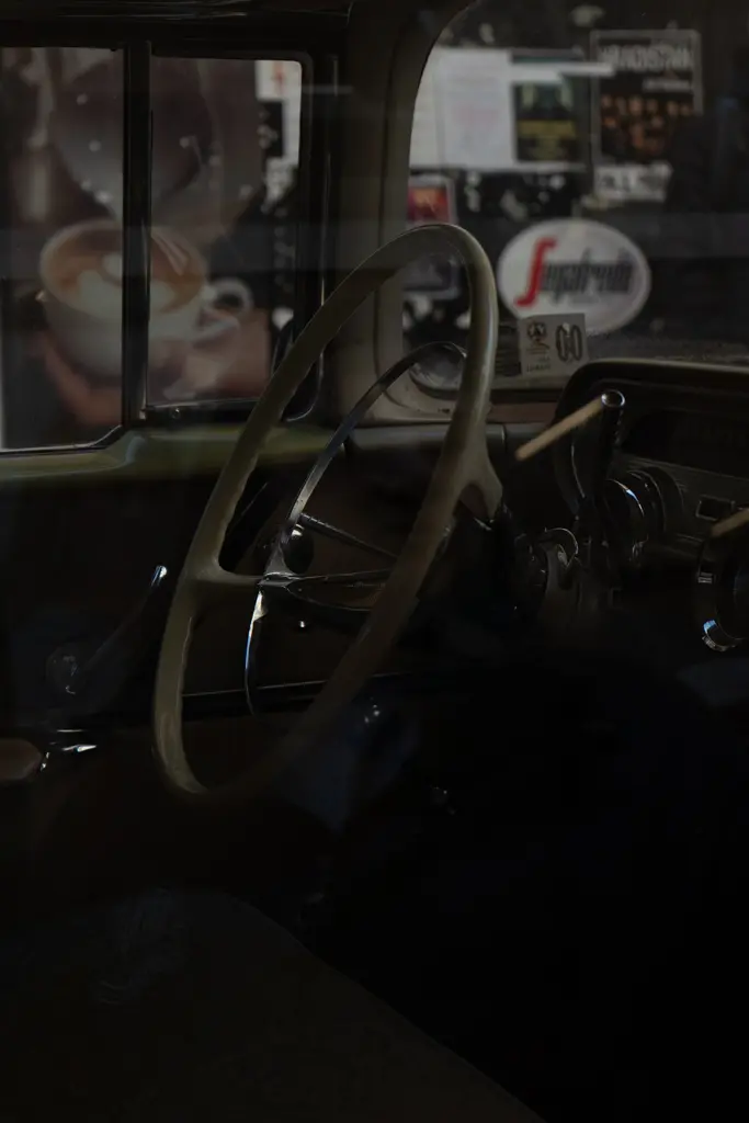 Pohled přes okno na interiér historického automobilu s volantem, palubní deskou a odrazy okolí ve skle.